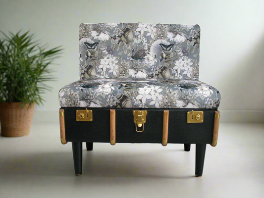 A chair made from a vintage steamer trunk with a floral fabric cover, black frame, gold painted fittings, and a varnished finish.